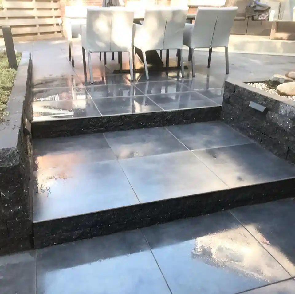 Brede trap van antracieten tegels met opstanden van zwarte stapelblokken tussen twee terrasniveaus, met een eethoek met lichtgrijze stoelen boven aan de trap