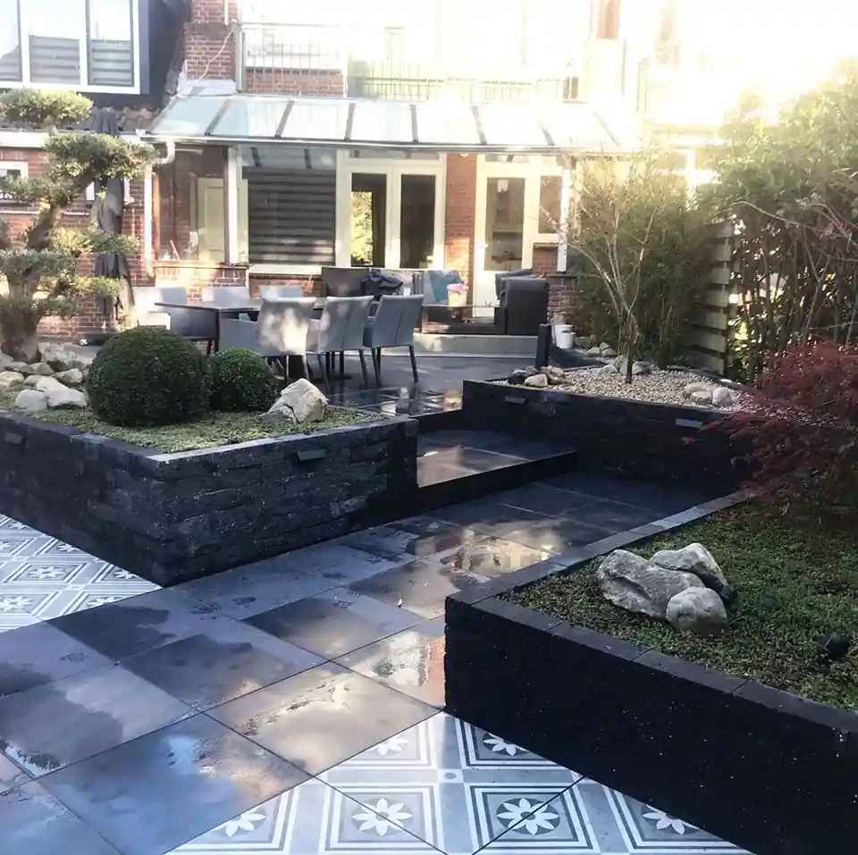 Verhoogde borders van zwarte stapelblokken met sedum en zwerfkeien rond een brede trap, met een terras van patroontegels en antracieten tegels