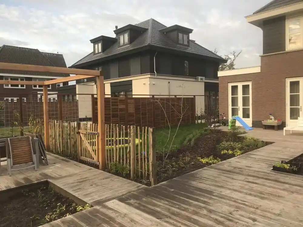 Houten pergola naast een schapenhek met poort dat een speelhoek met gazon afschermt, met lattenschermen langs de erfgrens en een brede vlonder