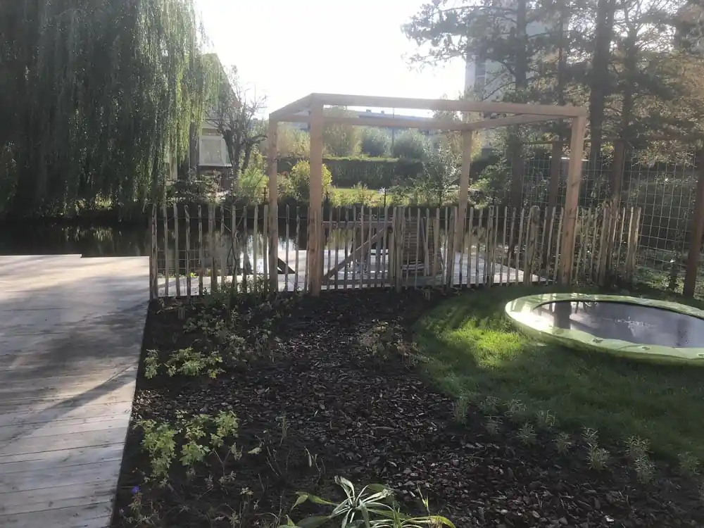 Houten pergola bij een schapenhek aan het water, met een verdiepte trampoline in het gazon en een border met schors en jonge beplanting