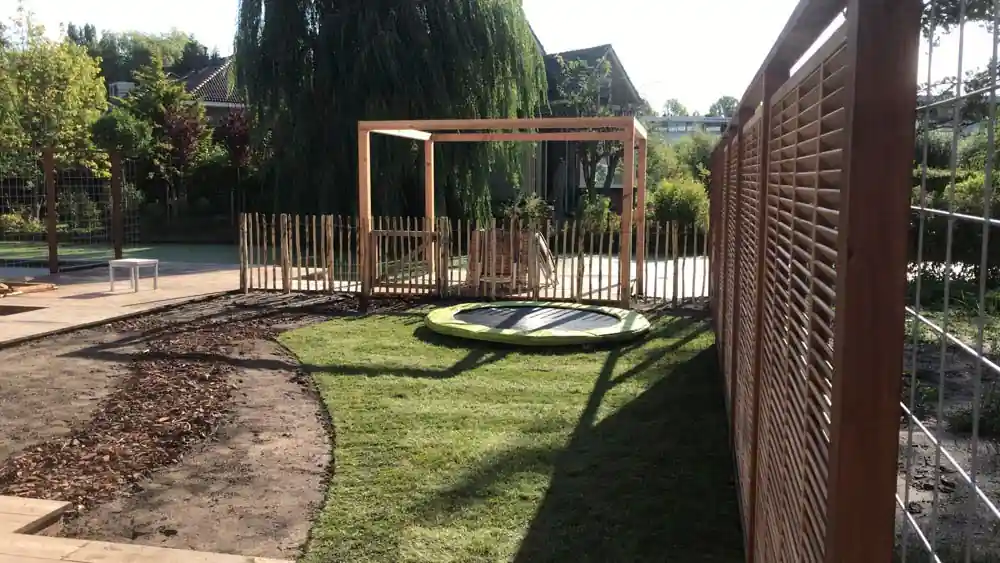 Vers gelegd gazon met verdiepte trampoline langs een hoog lattenscherm, met een schapenhek, pergola en een border met boomschors