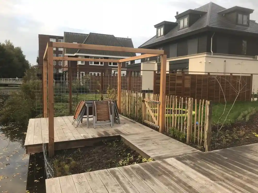 Houten vlonder met pergola en tuinset direct aan de waterkant, met een schapenhek en gaaspanelen als afscheiding naar het naastgelegen perceel