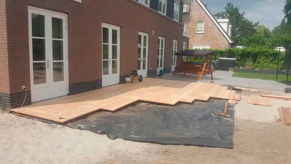 Houten vlonder in aanbouw langs de gevel met openslaande deuren, met losse planken en worteldoek op het zand ernaast