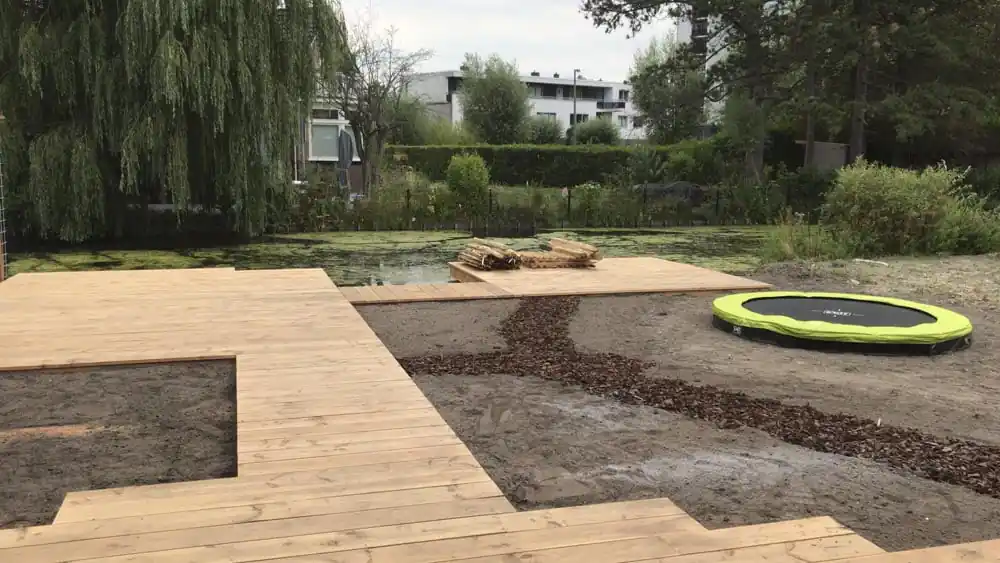 Brede houten vlonder met uitsparingen voor beplanting langs de slootkant, met een groene verdiepte trampoline en aangelegde schorspaden in het zand