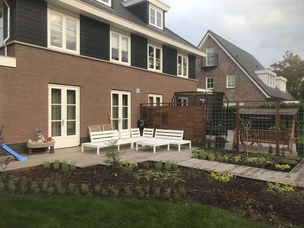 Ruime houten vlonder met witte loungebanken langs de gevel van een woning met openslaande deuren, met verse borders en een gazon op de voorgrond