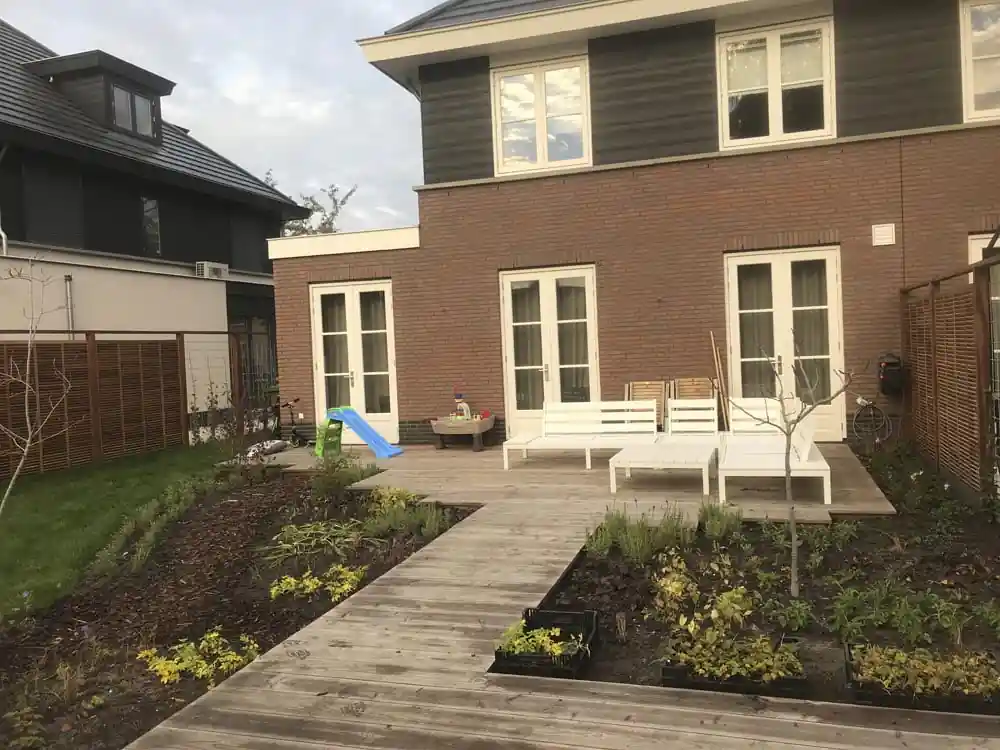 Houten vlonderpad dat door verse borders naar het terras met witte loungebanken loopt, met een jonge boom en een kinderglijbaan bij de woning