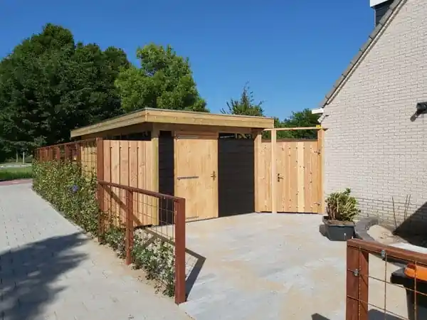 Houten berging met plat dak en een dubbele poort naast de zijgevel, met een bestrate oprit en een hekwerk met klimplanten ernaast