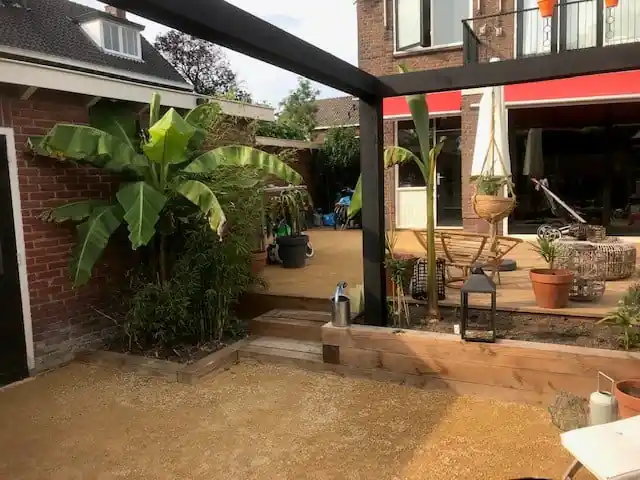 Verhoogde border van houten balken met bananenplanten en bamboe langs een vlak geel dolomiet, met een zwarte pergola en de vlonder met loungeset erachter