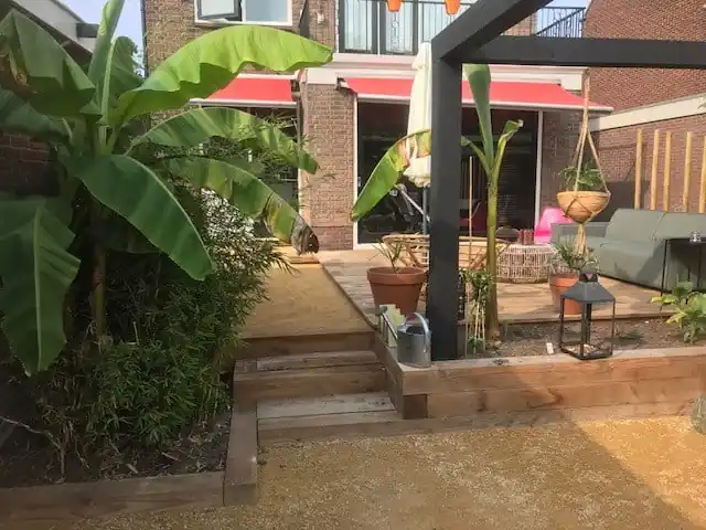 Brede houten traptreden die van de vlonder naar het lager gelegen dolomiet leiden, met bananenplanten in de border en een zwarte pergola erboven