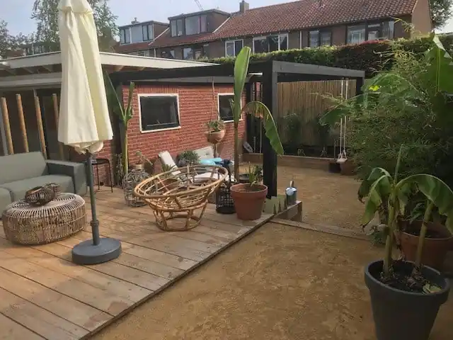 Overzicht met een houten vlonder met loungeset en parasol, een zwarte pergola met schommel boven het dolomiet en bananenplanten in potten en borders