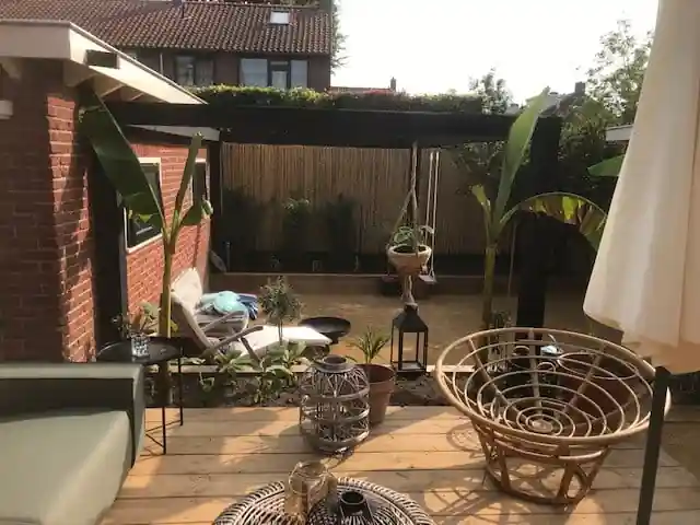 Ronde rotan fauteuil en lantaarns op een houten vlonder, met een ligstoel tegen de bakstenen gevel en een zwarte pergola boven het dolomiet