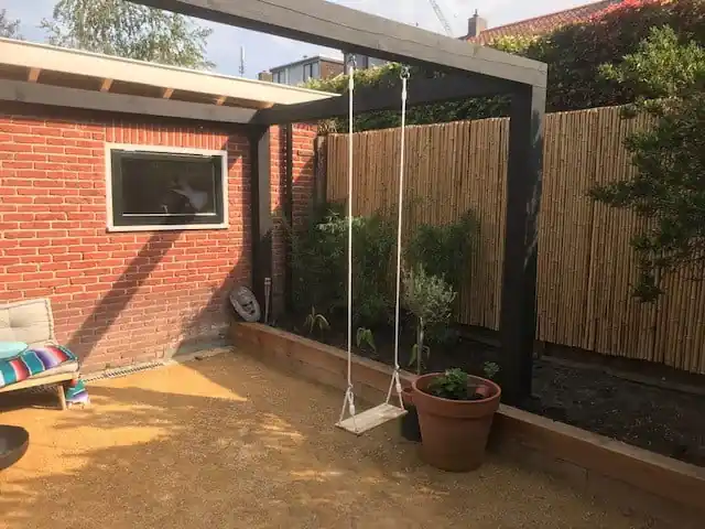 Houten schommel aan een zwarte pergola boven een vlak geel dolomiet, met een bamboeschutting, een olijfboom in terracotta pot en een border met houten keerwand