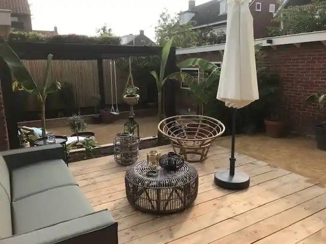 Houten vlonder met loungebank, rotan fauteuil en parasol, grenzend aan een vlak geel dolomiet, met een zwarte pergola en bamboeschutting op de achtergrond