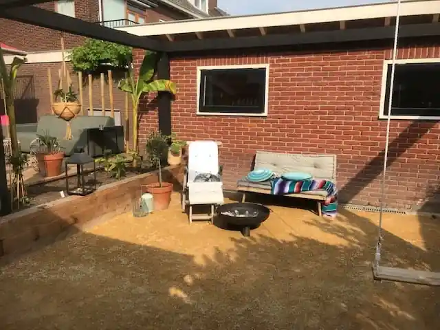 Zitbank met kussens en een ronde vuurschaal op een vlak geel dolomiet voor een rode bakstenen gevel, met bananenplanten in een verhoogde border van houten balken