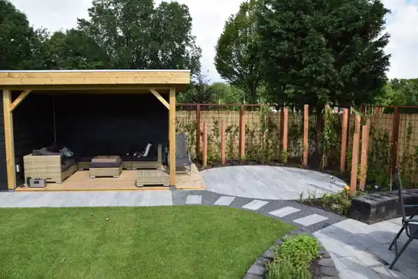 Houten overkapping met een rieten loungeset op een vlonder, met daarvoor een gazon, een terras met stapstenen en een border langs de schutting