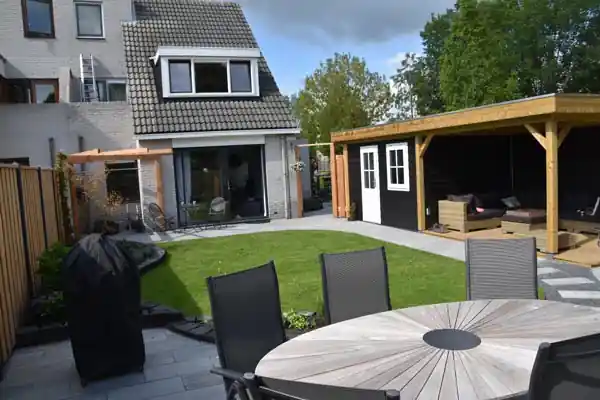 Achtertuin met een houten overkapping met loungeset naast een donker tuinhuis met witte deuren, met gazon en een terras met eettafel op de voorgrond