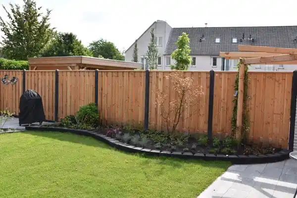 Gebogen border met jonge beplanting en een boompje langs een houten schutting, afgezet met zwarte banden, grenzend aan een gazon