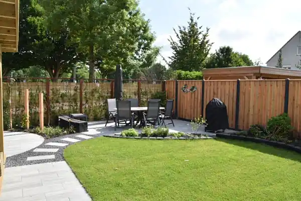 Terras met een tuinset en parasol naast een rond gazon, omsloten door een houten schutting met klimplanten en een border met lage beplanting