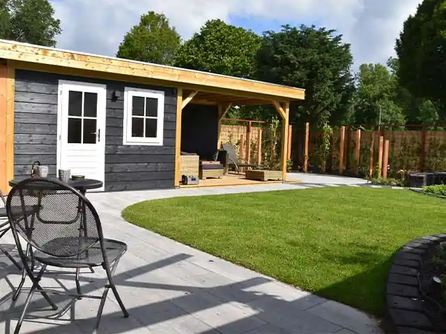 Donker houten tuinhuis met aangebouwde overkapping boven een houten vlonder met loungeset, met daarnaast een terras en een rond gazon