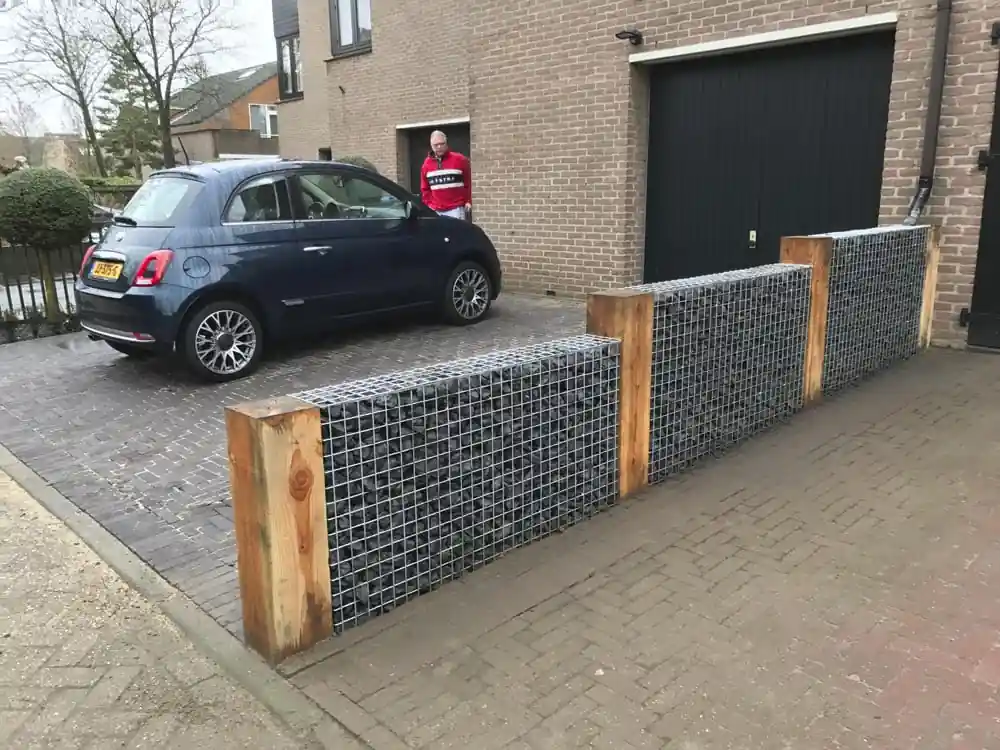 Lage erfafscheiding van schanskorven gevuld met grijze breuksteen, afgewisseld met houten staanders, langs een bestrate oprit met geparkeerde auto