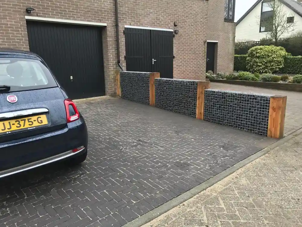 Bestrate oprit van donkere klinkers in keperverband langs een rij schanskorven met grijze breuksteen tussen houten staanders, met een auto voor de garagedeur