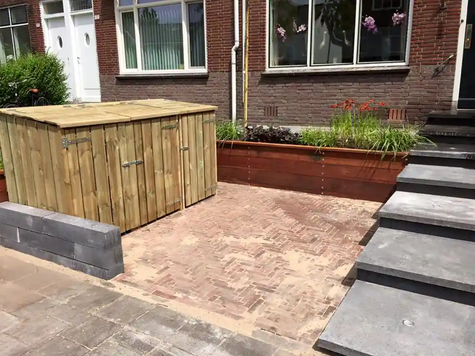 Houten containerberging met deksel naast een terras van rode klinkers in visgraatverband, met een hardhouten plantenbak met siergrassen langs de gevel