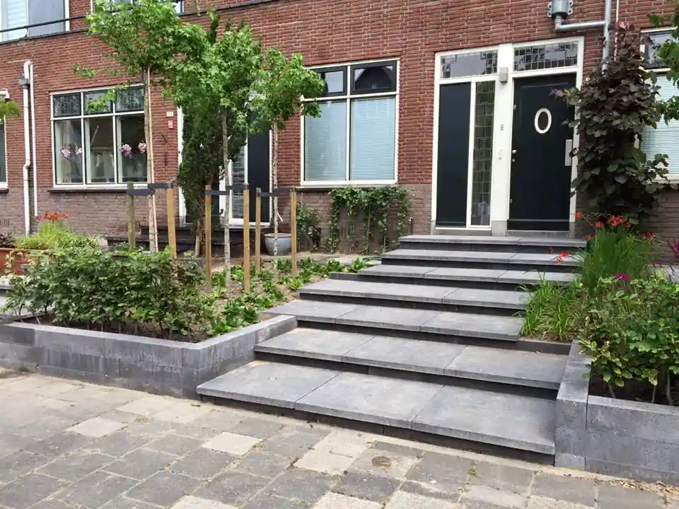 Trap van antracieten traptreden naar de voordeur, geflankeerd door verhoogde borders met jonge bomen op boompalen, beukenhaag en oranje montbretia