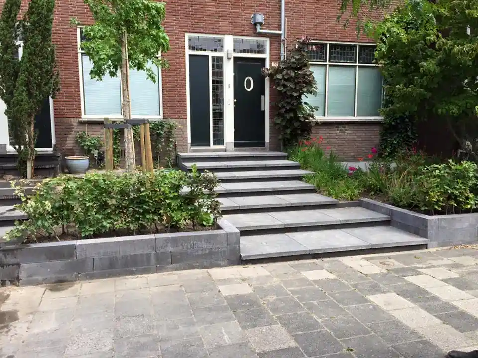 Brede trap van antracieten traptreden naar de donkere voordeur van een jarendertigwoning, tussen verhoogde borders van stapelblokken met beuk en montbretia