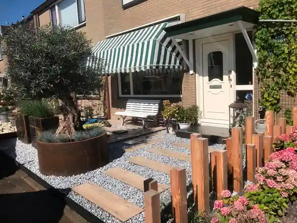 Voortuin met een ronde cortenstalen plantenbak met olijfboom, een pad van houten stapstenen in wit split en een rij Douglas paaltjes als lage erfafscheiding