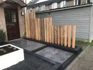 Detail van het scherm van losse Douglas balken op verschillende hoogtes, met daarvoor een terras van grote grijze tegels en een strook zwart split
