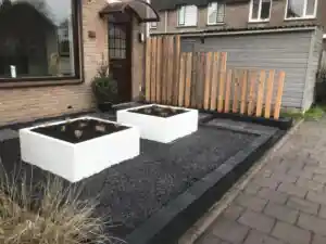 Twee witte plantenbakken op een vlak zwart split, met een scherm van losse Douglas balken als erfafscheiding en een antracieten kantopsluiting langs de stoep