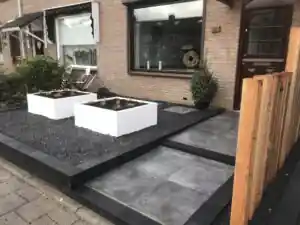 Pad van grote grijze tegels naar de voordeur, langs witte plantenbakken op zwart split, met een buxus in pot en het Douglas scherm op de voorgrond
