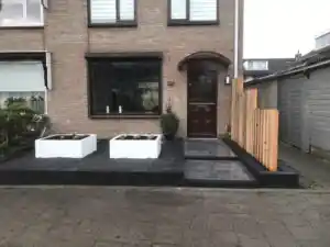 Vooraanzicht van de lichte bakstenen woning met donkere voordeur, met twee witte plantenbakken op zwart split en een scherm van Douglas balken rechts