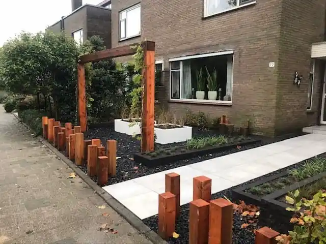 Houten pergola met klimplant boven een vlak zwart split, met witte plantenbakken en een rij houten paaltjes op verschillende hoogtes als erfafscheiding