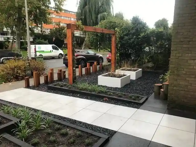 Pad van grote lichte tegels langs borders met siergrassen achter antracieten randen, met witte plantenbakken op zwart split onder een houten pergola