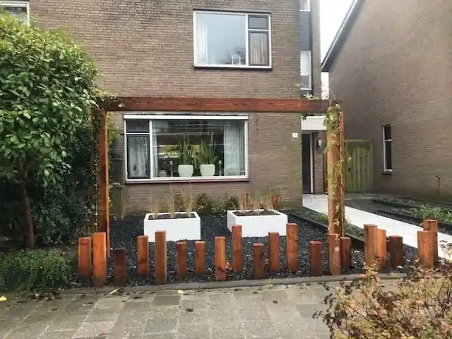 Vooraanzicht van de voortuin met een houten pergola langs de gevel, witte plantenbakken op zwart split en een rij houten paaltjes langs de stoep