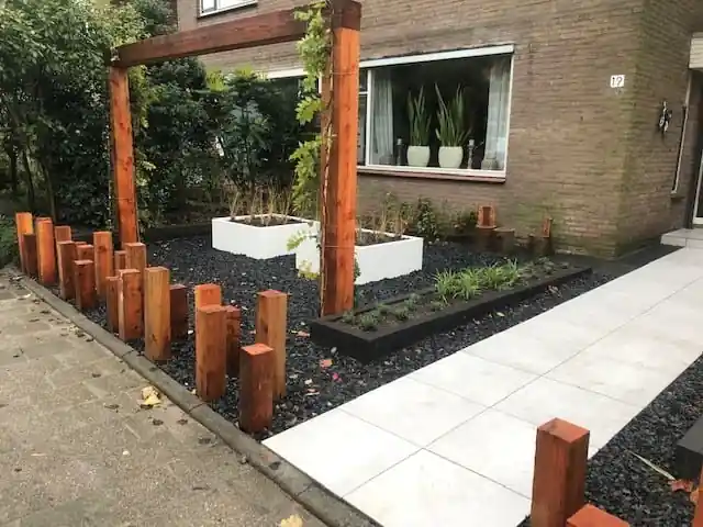 Twee witte plantenbakken met siergrassen op een vlak zwart split onder een houten pergola, met een border met jonge beplanting langs het tegelpad