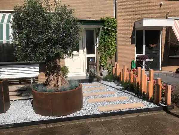 Pad van houten stapstenen door wit split naar de voordeur, langs een ronde cortenstalen bak met olijfboom en blauwe siergrassen en Douglas paaltjes