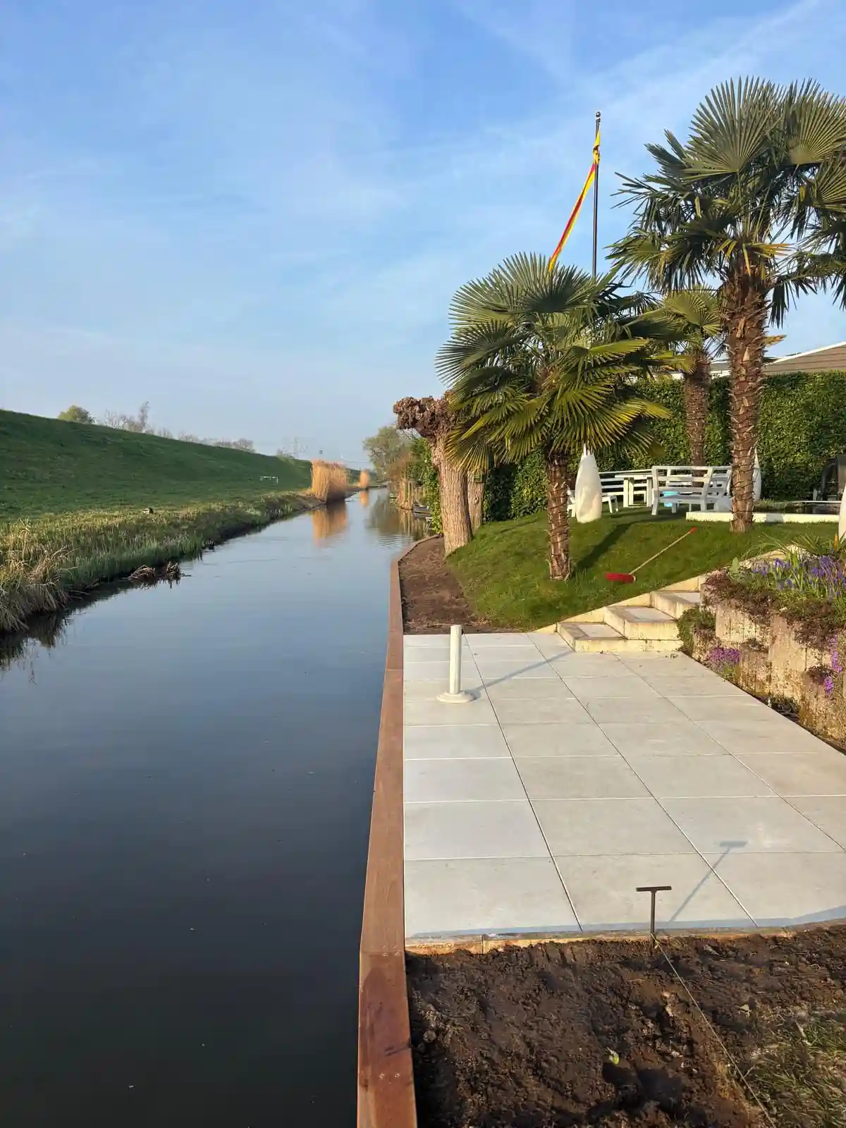 Strakke cortenstalen damwand langs de waterkant met daarachter een terras van lichte tegels, een gazon met palmbomen, een witte tuinbank en een vlaggenmast