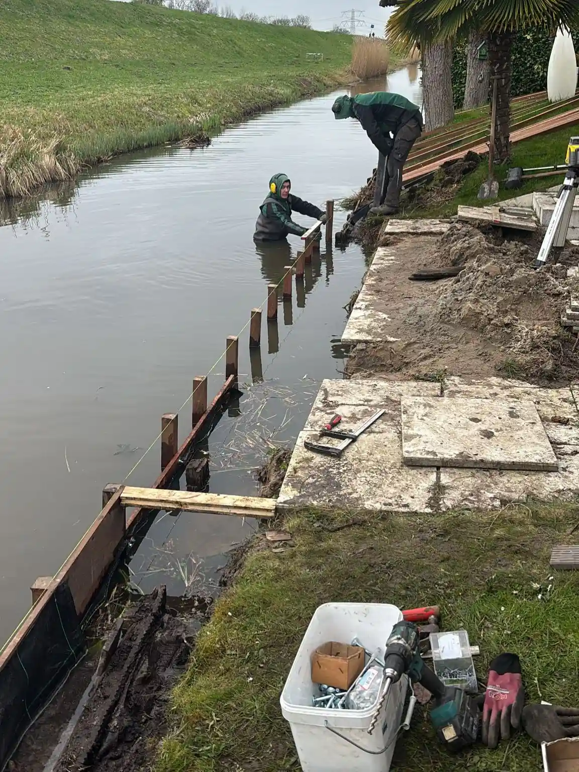 Twee hoveniers plaatsen een stalen damwand langs de oever, een van hen staat in waadpak in het water terwijl de ander de planken aanreikt, met gereedschap op de kant