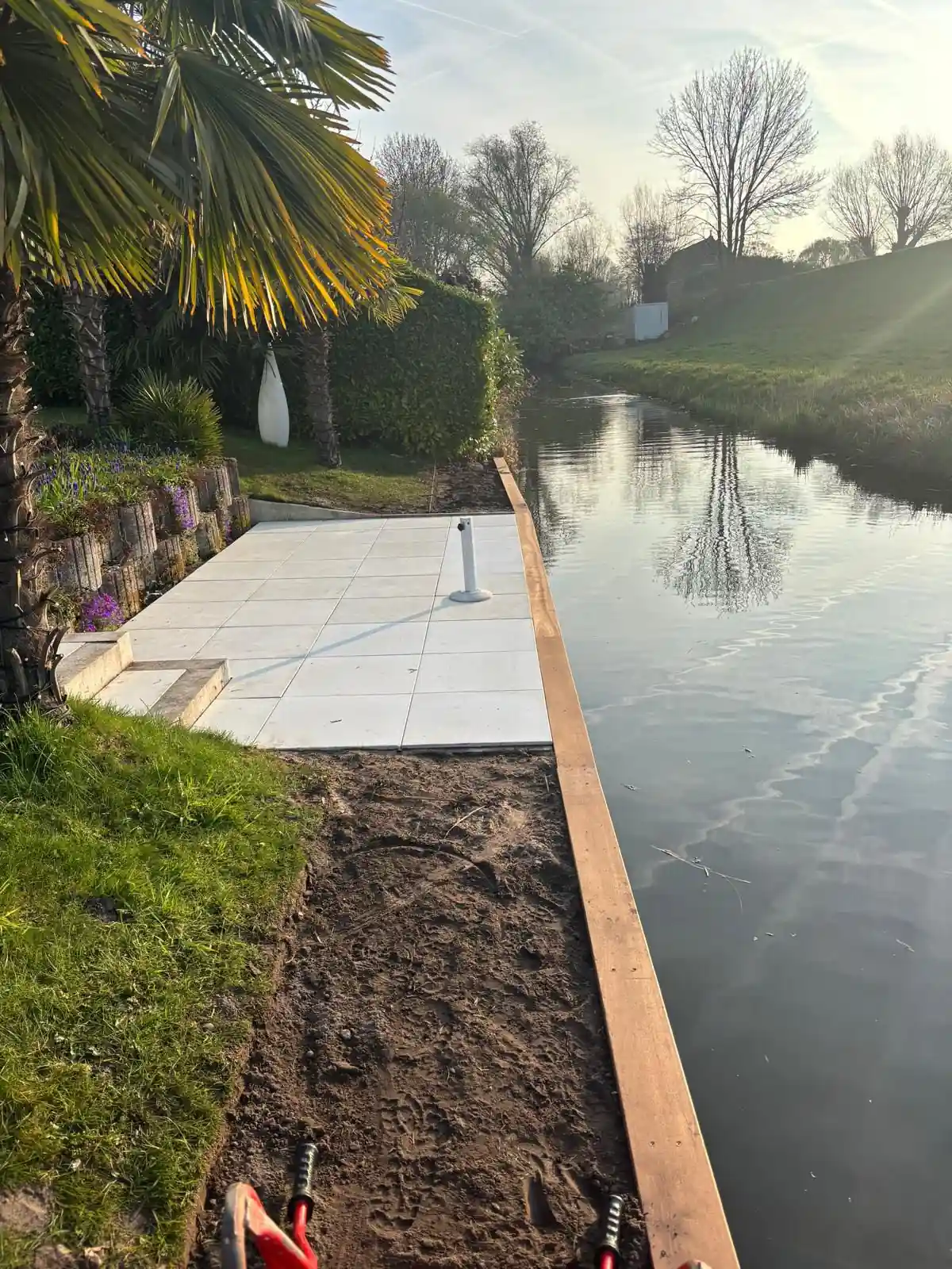 Terras van lichte tegels direct aan de oever, afgezet met een cortenstalen damwand langs het water, met palmen en een border met paarse voorjaarsbloeiers erachter