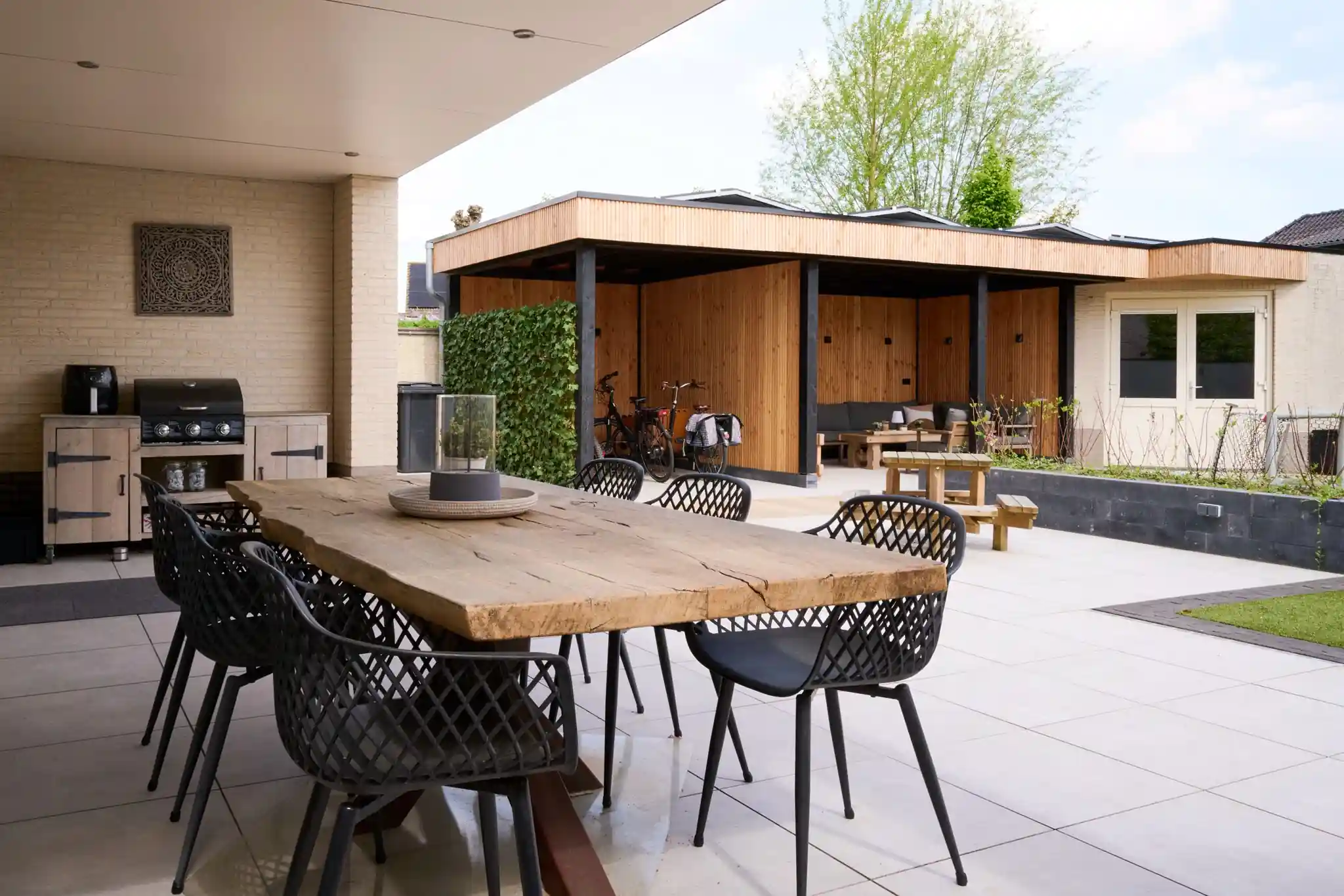 Robuuste houten eettafel met zwarte kuipstoelen onder het afdak bij een buitenkeuken met barbecue, met zicht op de tuinkamer en een klimopwand