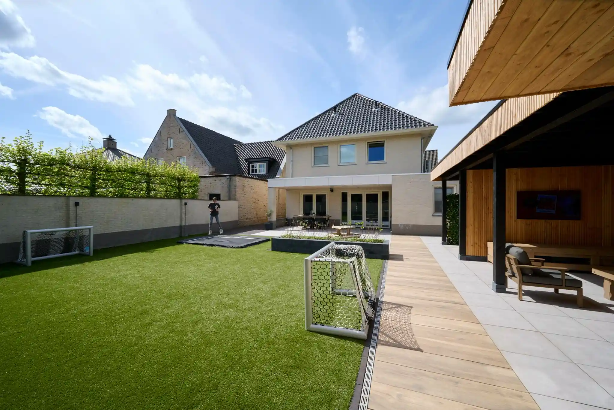 Groot veld kunstgras met twee voetbaldoelen en een verdiepte trampoline, omsloten door een lichte muur met leihaag, met de woning en de tuinkamer erachter