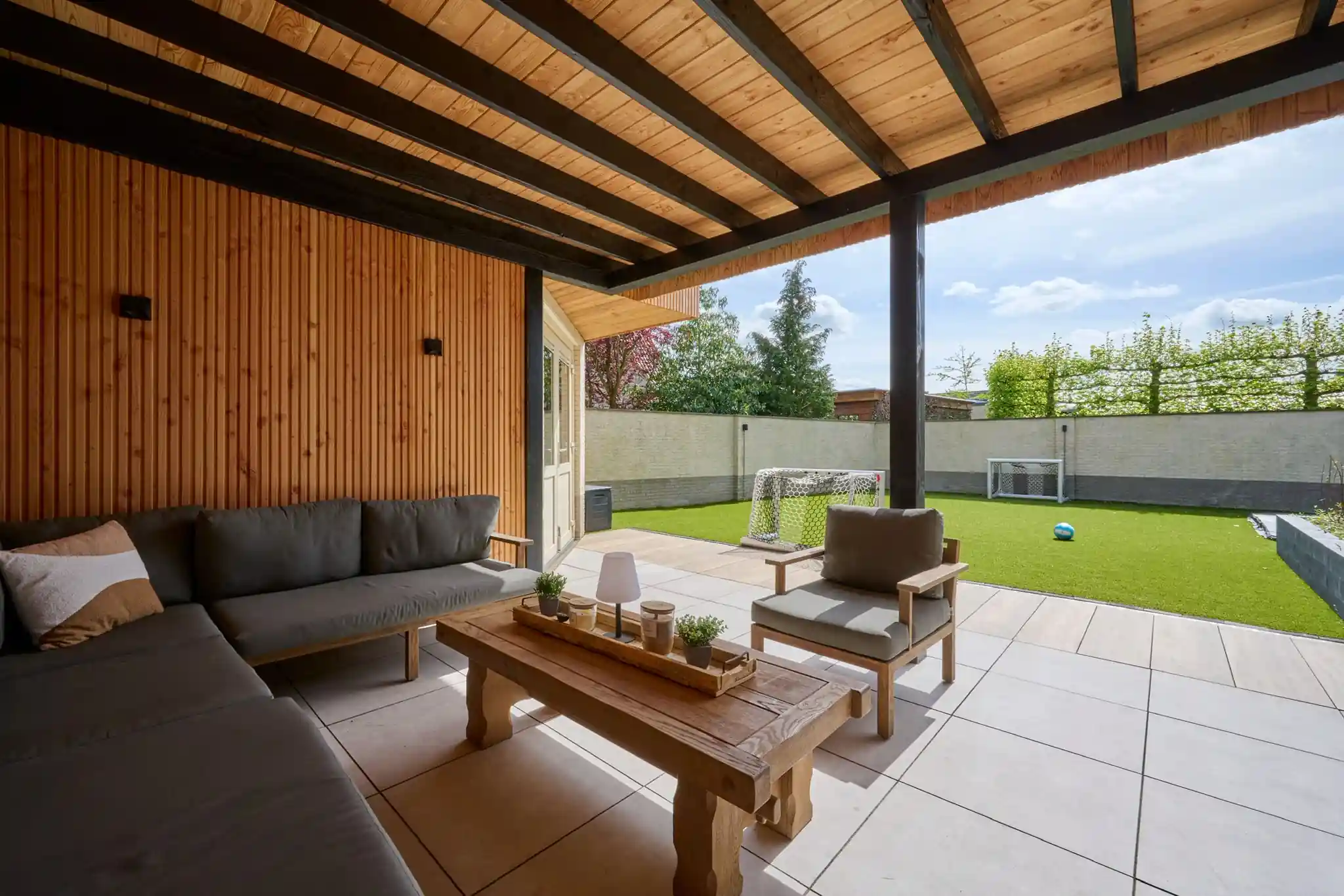 Loungehoek met grijze hoekbank en houten salontafel onder een overkapping met zwarte balken, met uitzicht op het kunstgras met voetbaldoelen