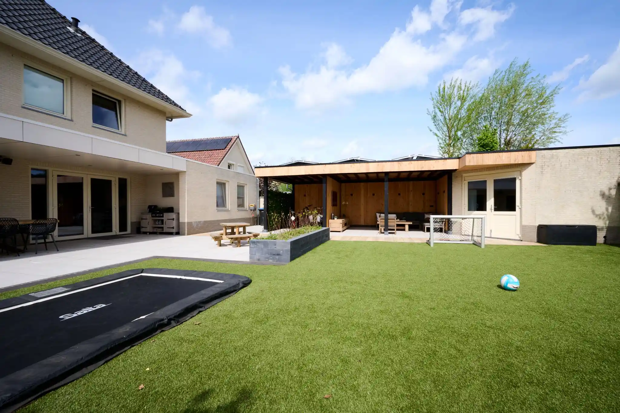 Ruim veld kunstgras met verdiepte trampoline en voetbaldoel, met een tuinkamer met Douglas wanden, een verhoogde border van stapelblokken en een picknicktafel