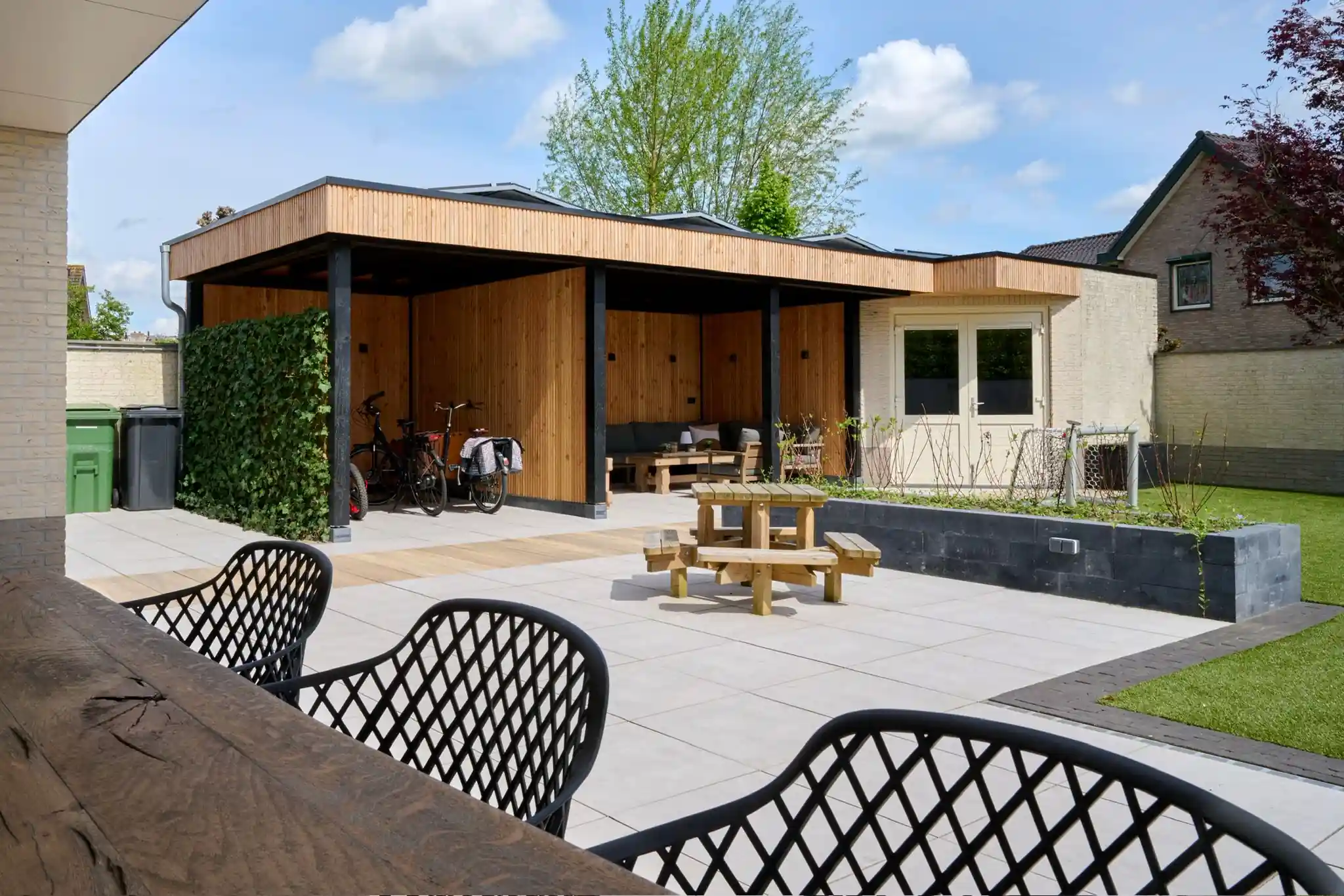 Ruim terras van grote lichte tegels met een houten picknicktafel, voor een tuinkamer met Douglas betimmering en een klimopwand bij de fietsenstalling