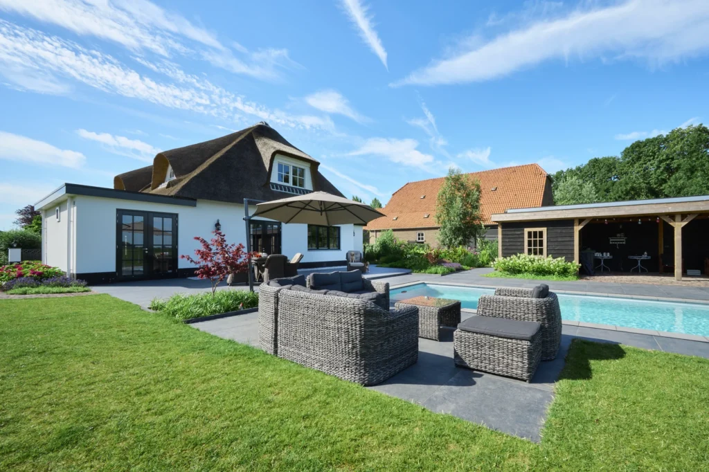 Achtertuin met zwembad, een rieten loungeset met parasol op een terras van grote grijze tegels, gazon en een overkapping bij een zwarte tuinberging