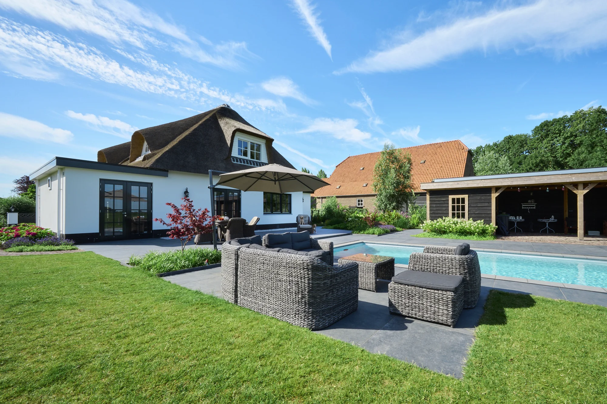 Achtertuin met zwembad, een rieten loungeset met parasol op een terras van grote grijze tegels, gazon en een overkapping bij een zwarte tuinberging