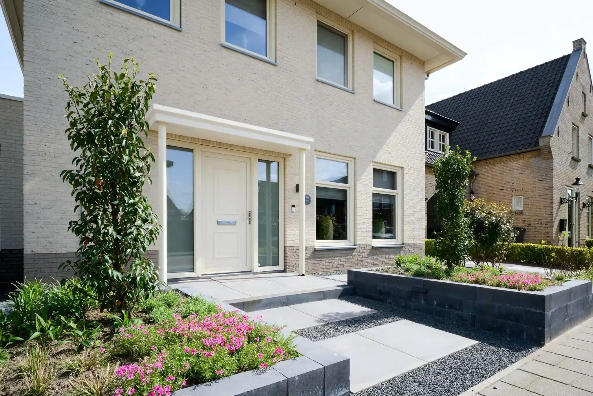 Entree met lichte voordeur en een pad van grote tegels met grindvoegen, tussen verhoogde borders van antracieten stapelblokken met roze bloeiende vaste planten