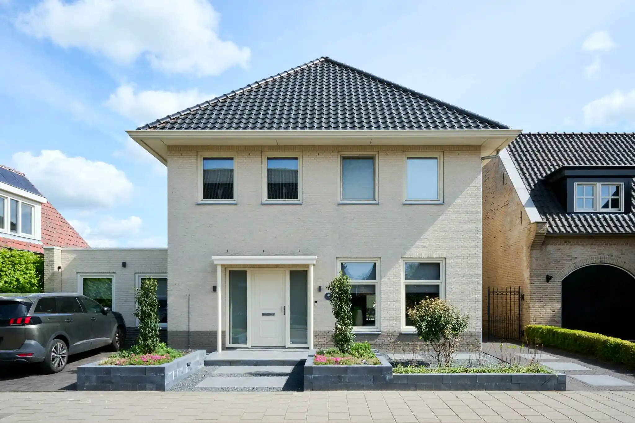 Vooraanzicht van de lichte bakstenen woning met twee verhoogde borders van stapelblokken, een tegelpad naar de voordeur en een bestrate oprit met auto
